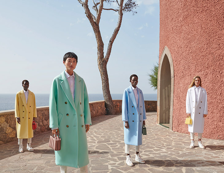 Four models wearing pastel Max Mara 101801 Light coats in a sunlit Mediterranean setting.