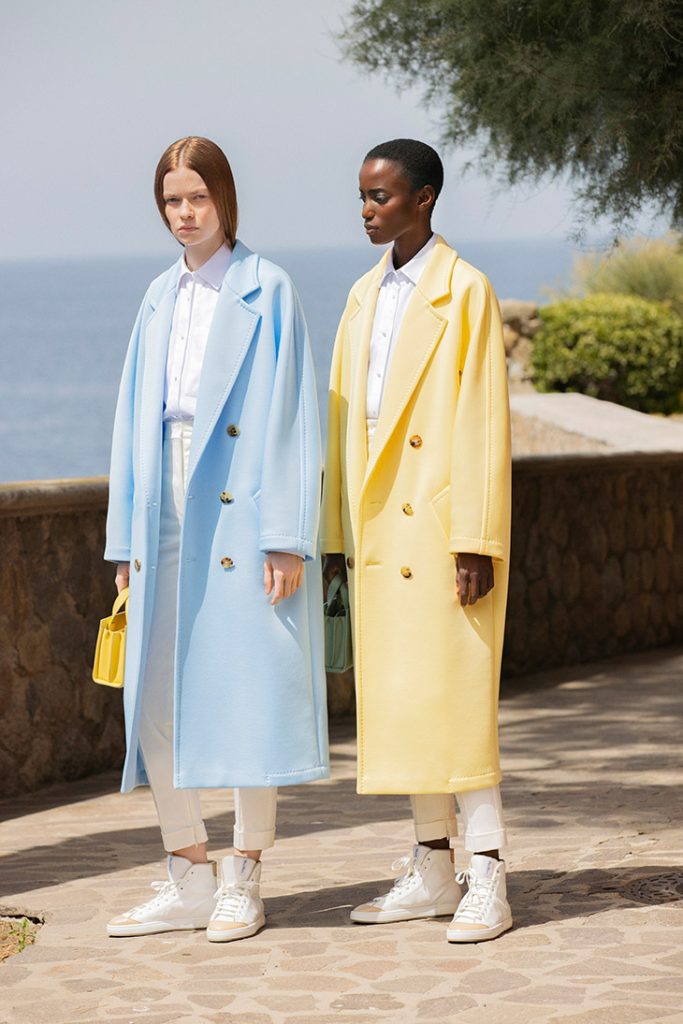 Two models wearing blue and yellow Max Mara 101801 Light coats in a Mediterranean setting.