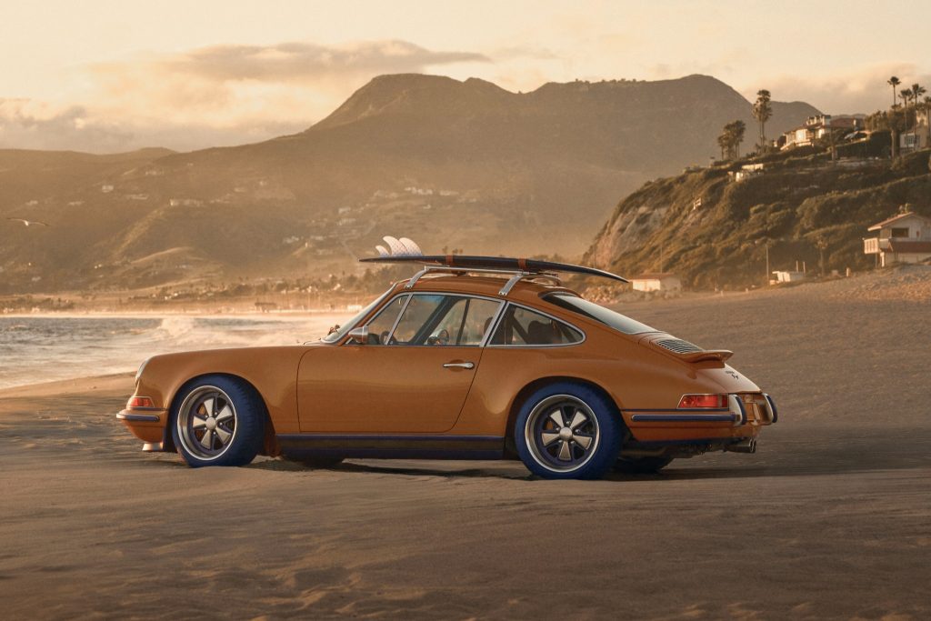 Saffron Porsche 911 Reimagined by Singer Classic on a beach at sunset with a surfboard mounted to its roof rack.