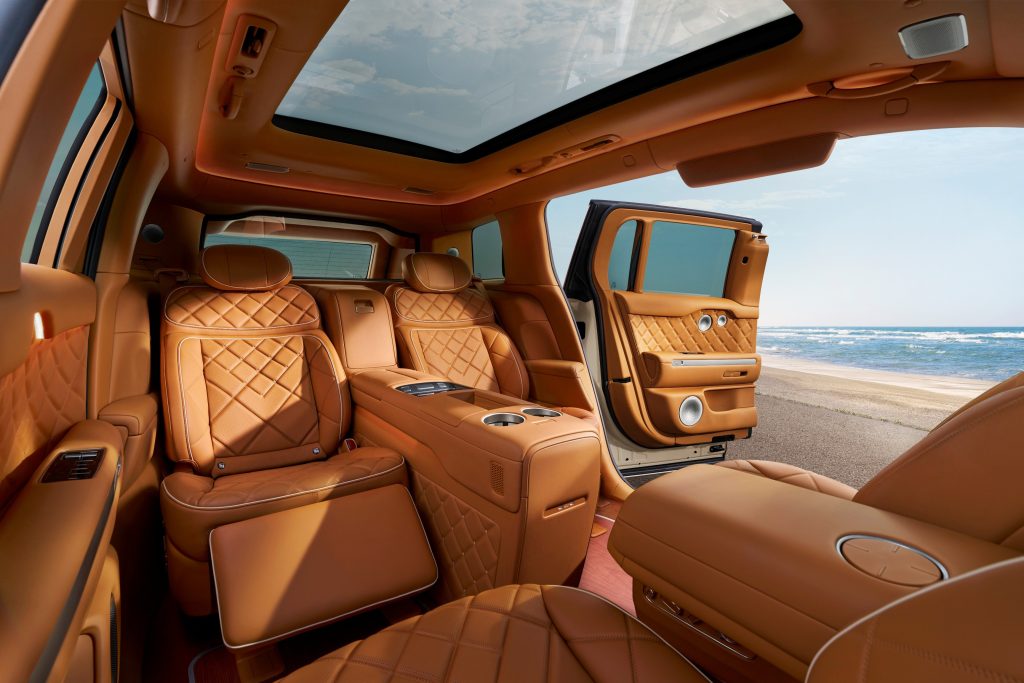 Tan executive second-row lounge inside the Genesis GV90 with an open coach door and sea view.