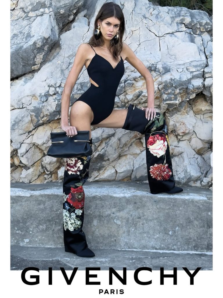 Givenchy Winter 2026: black cut-out bodysuit, floral over-the-knee boots and Pinch clutch against pale coastal rock.