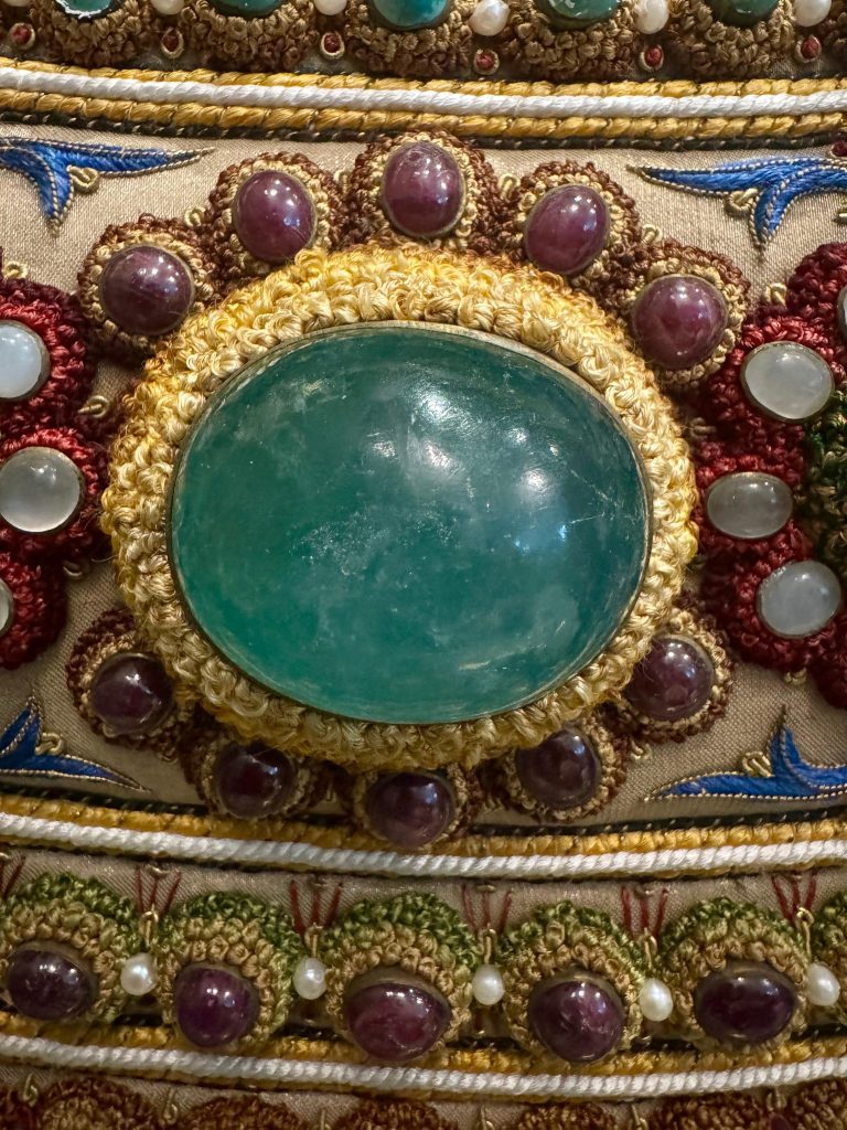 Emerald cabochon surrounded by gemstones and intricate embroidery in The Vase.