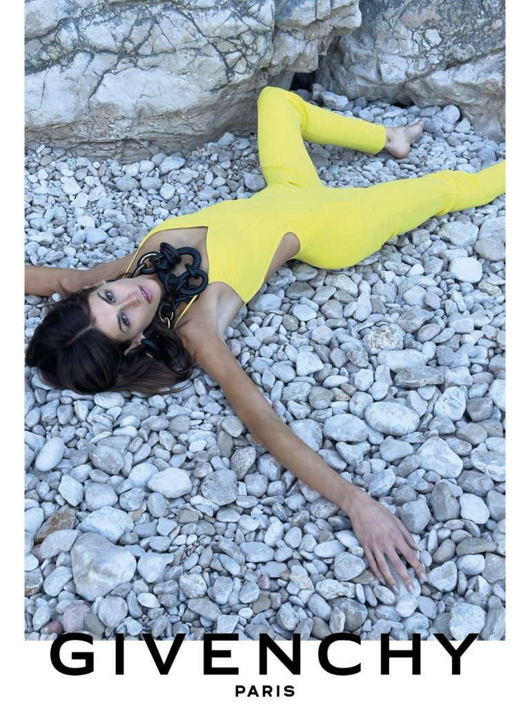 Model reclining on pale pebbles in a bright yellow cut-out all-in-one with an oversized black chain-link necklace.