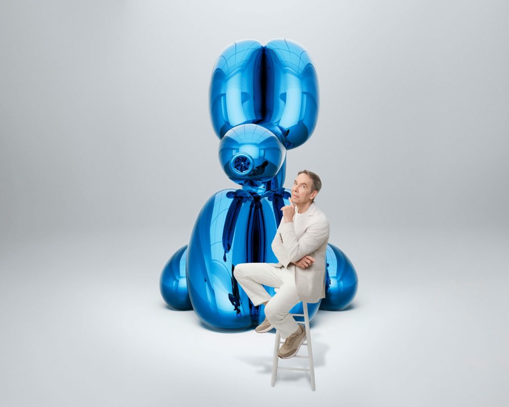 Jeff Koons seated beside a monumental blue Balloon Dog sculpture.