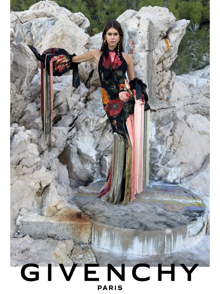 Model in a dark floral halterneck gown with long multicoloured fringe and voluminous matching arm pieces against coastal rock.