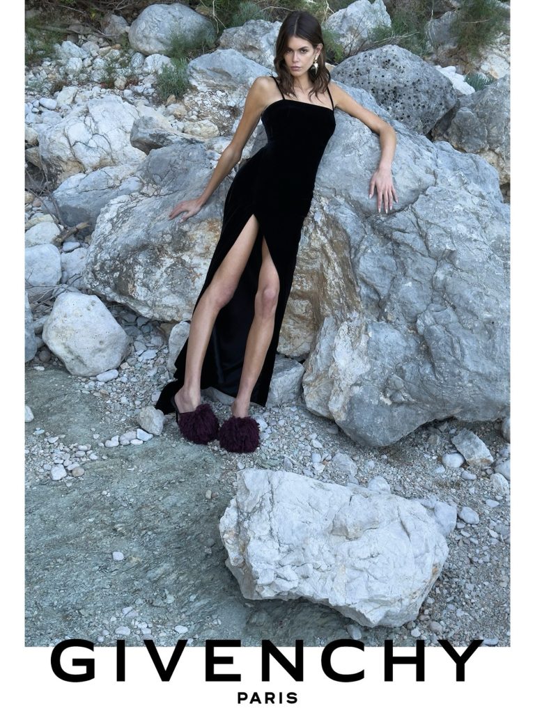 Model wearing a long black dress with thin straps, a high slit and dark plum pom-pom mules among pale rocks.